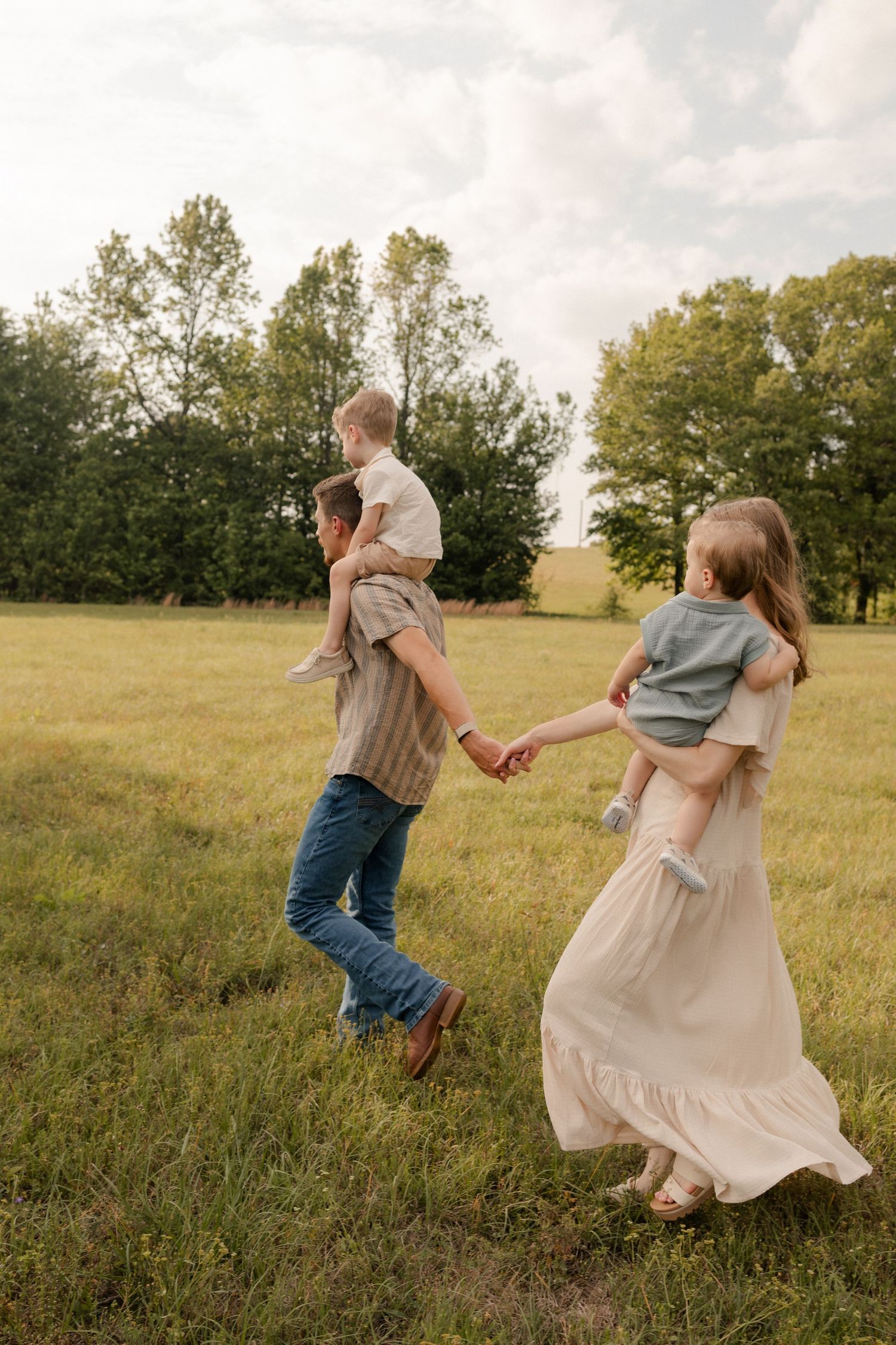 A family walks hand-in-hand through a green meadow, parents carrying young children on their backs and arms.