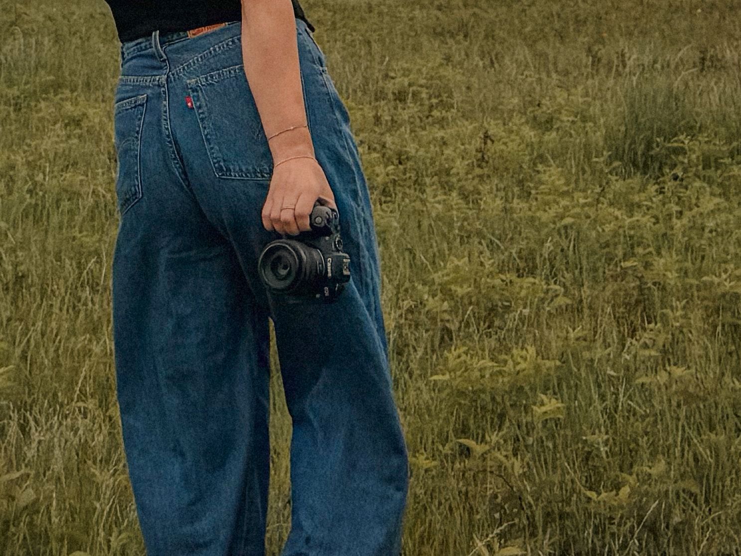 Close-up of woman in wide-leg denim jeans holding a black camera while standing in a lush green field.