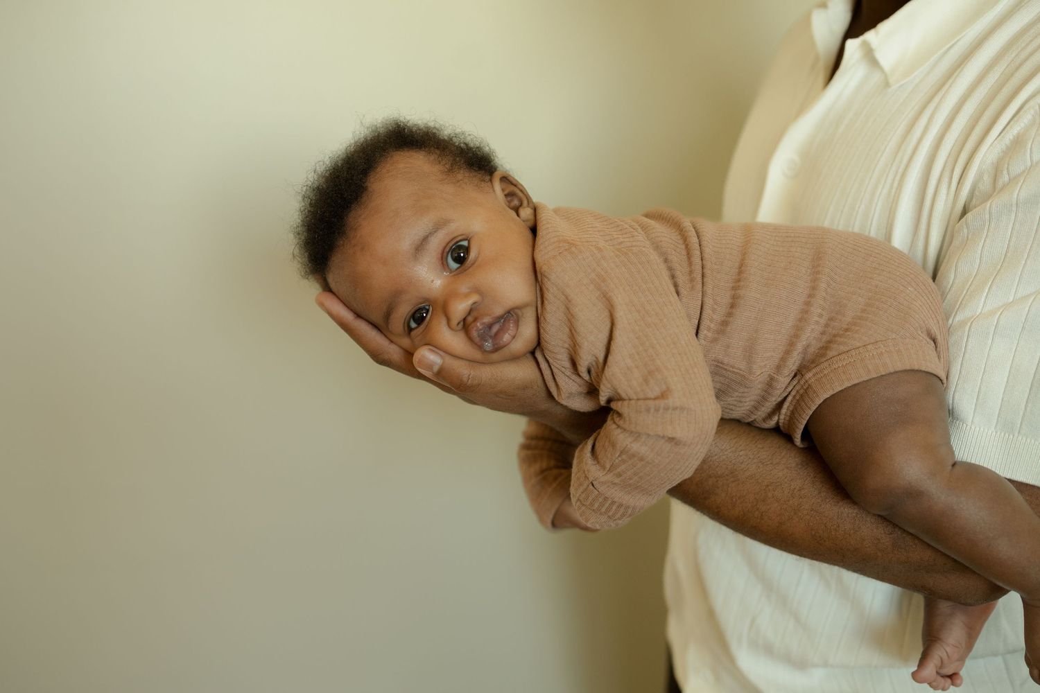 Black father cradling newborn baby in one hand against neutral wall background.