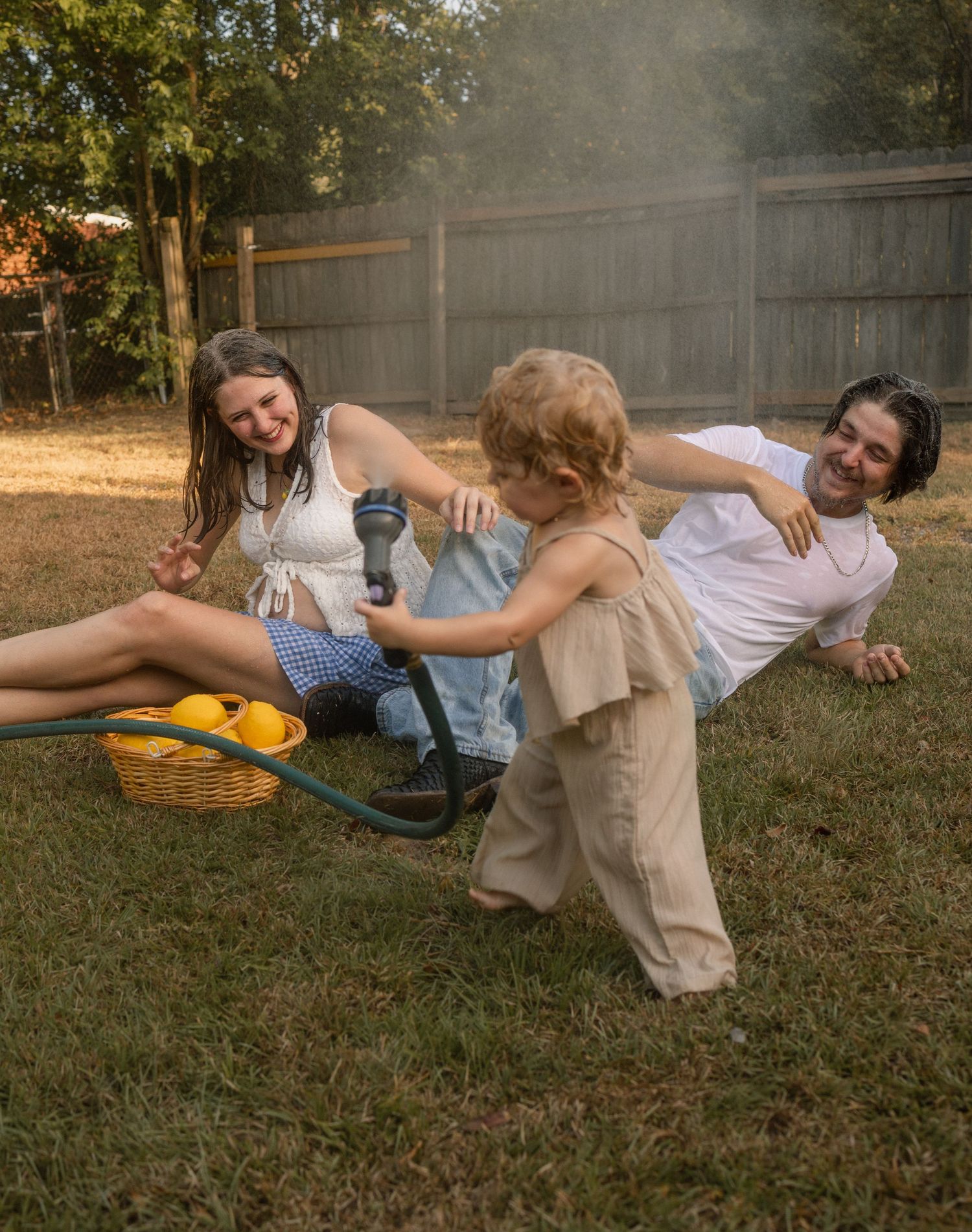 Family relaxing in backyard with toddler playing with garden hose near a basket of lemons.