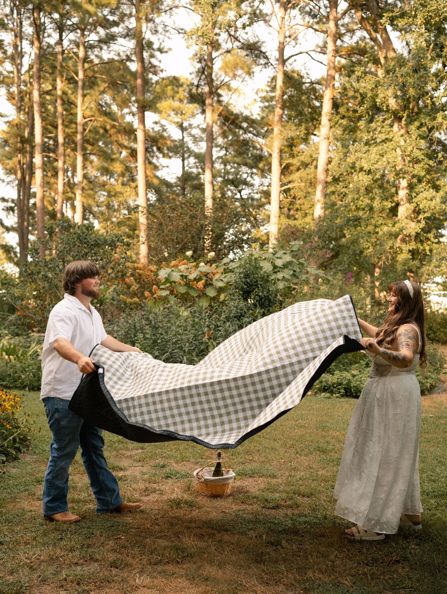 A couple spreads a gingham picnic blanket in a sunny garden surrounded by tall pine trees.