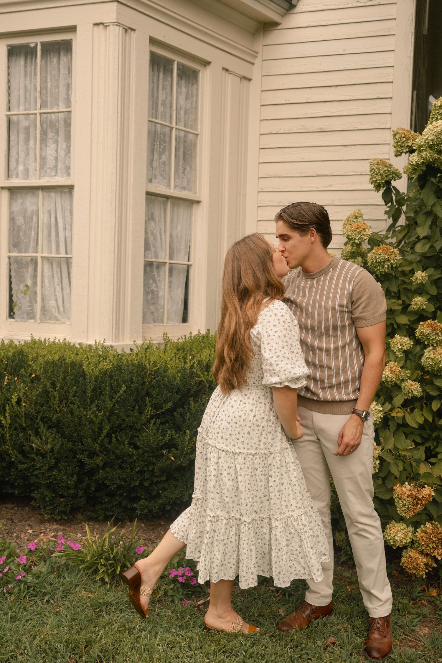 Couple sharing a sweet kiss beside a charming white house with hydrangea blooms in Cameron NC.