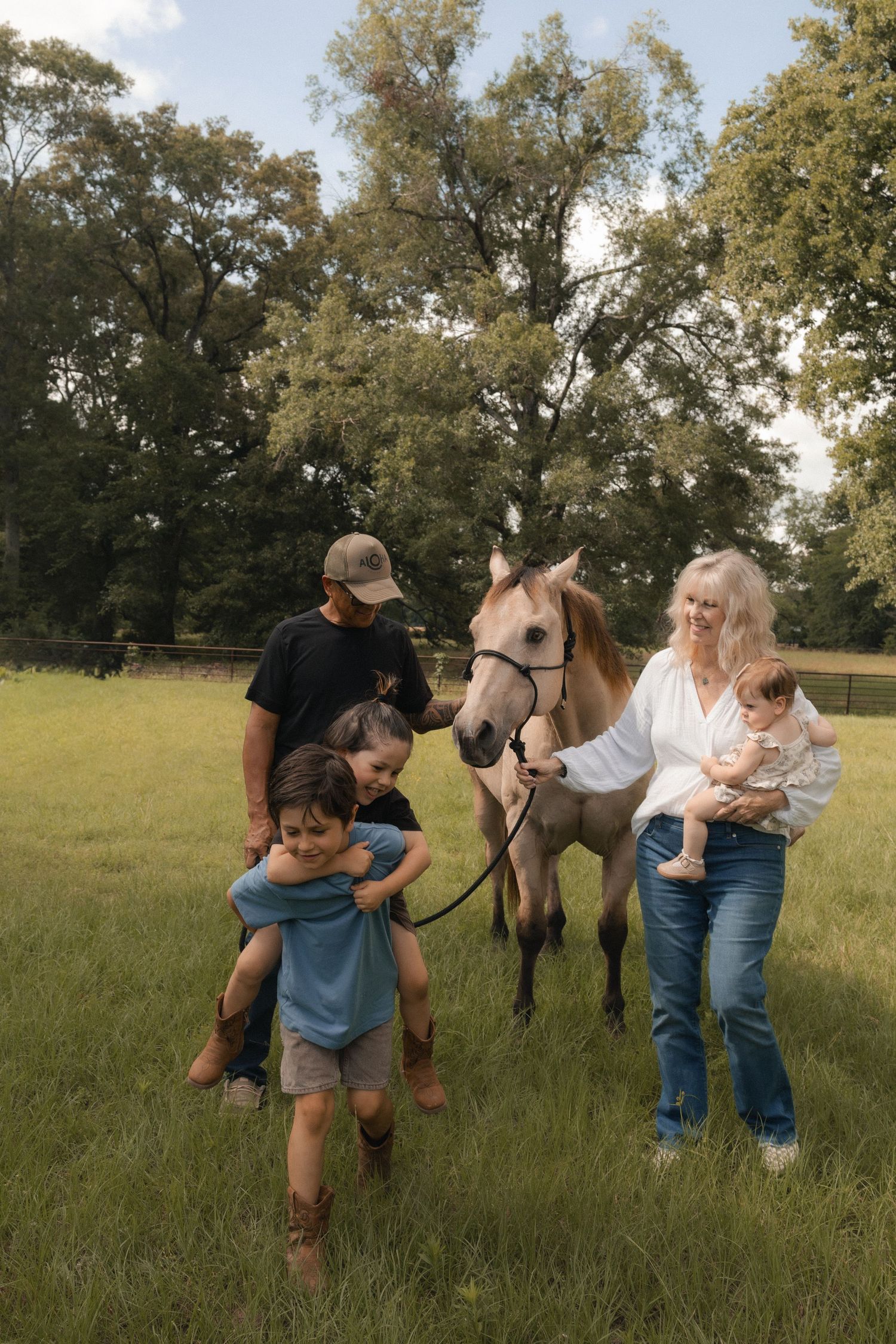 Family with horse on green farm, two children playing, woman holding baby, man in cap, surrounded by large trees.
