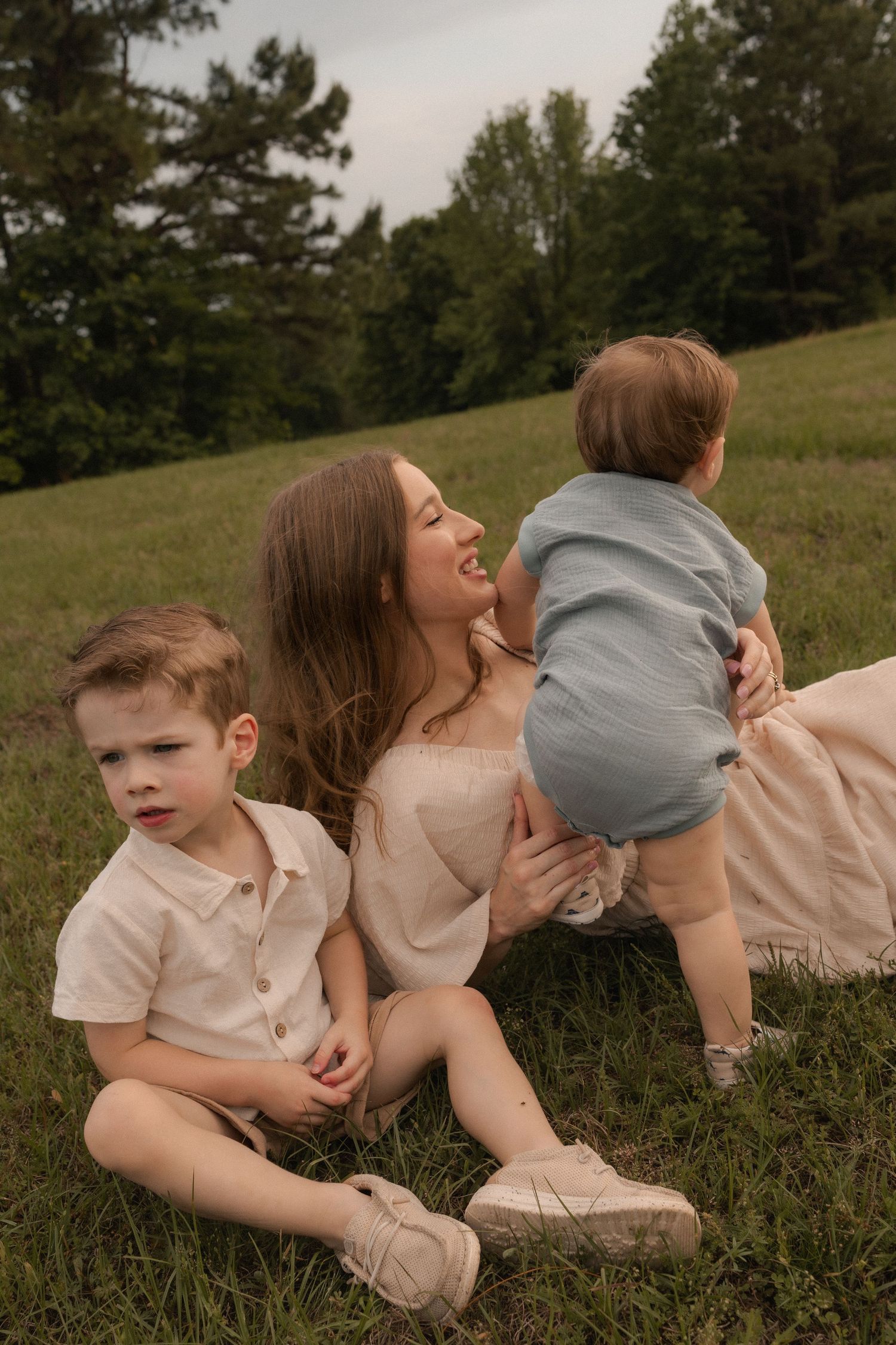 Mother sitting on grass with two young boys, one toddler standing and one child seated, in a lush green park.