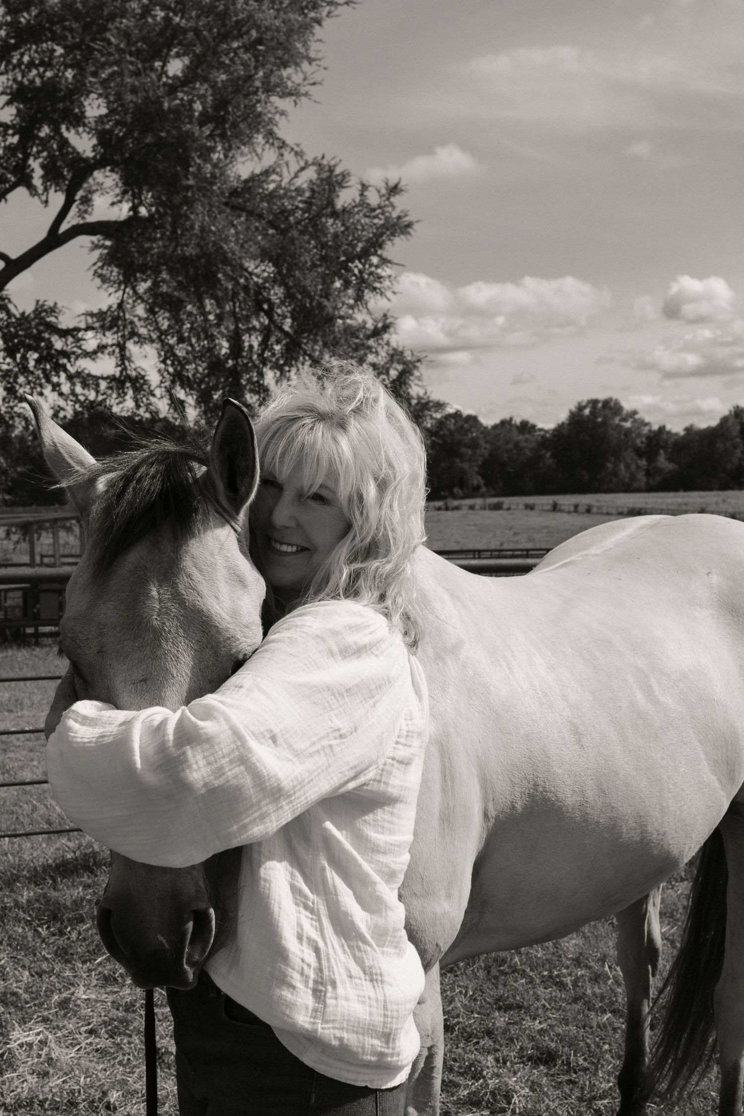 Smiling blonde woman embracing a white horse in a black and white farm photo.