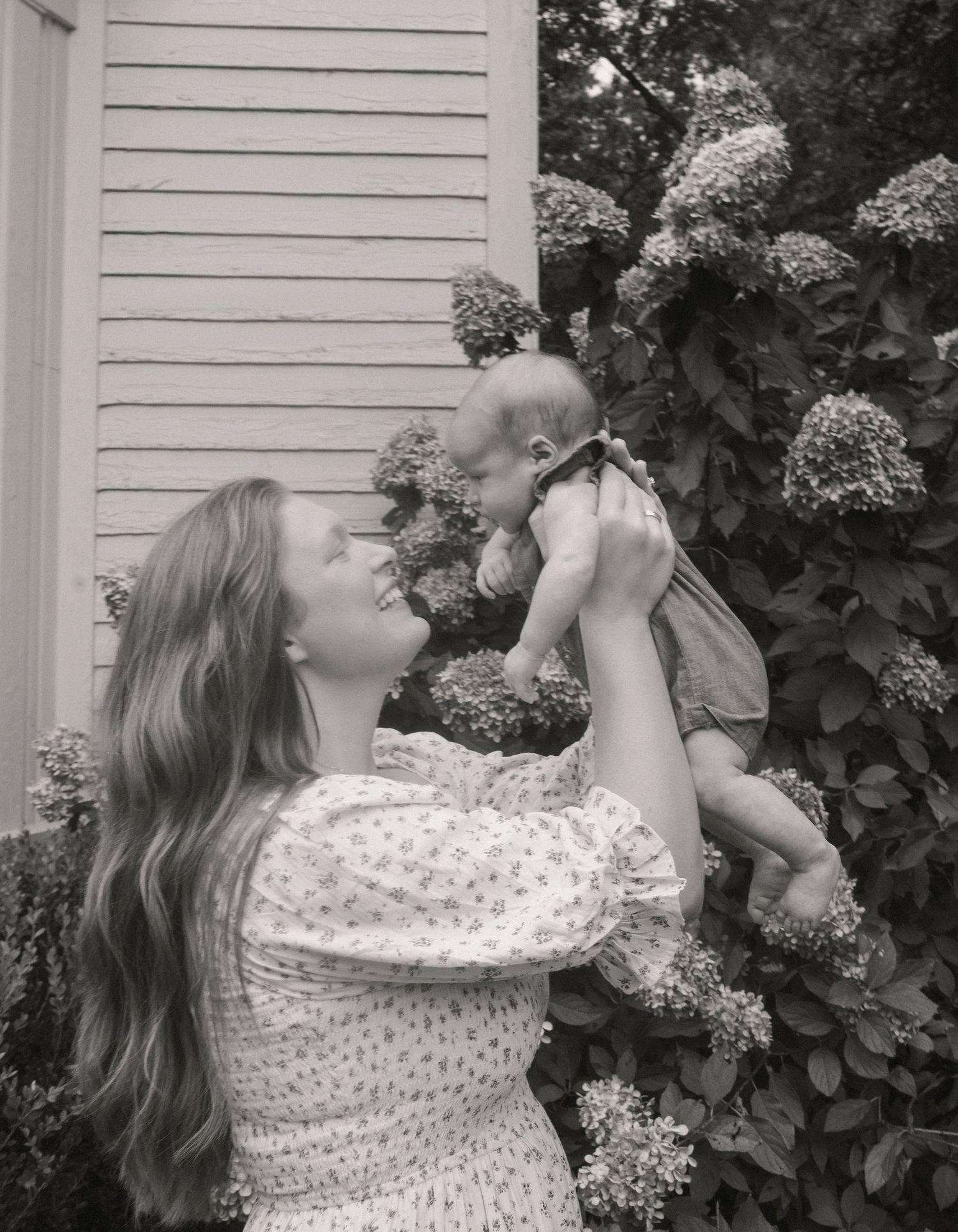 Black and white photo of mother lifting smiling baby near blooming hydrangea bush.