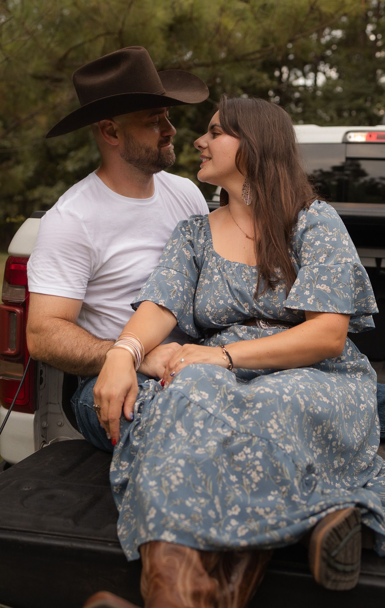 A couple sits intimately on a truck tailgate, man in cowboy hat and woman in blue floral dress gazing at each other.