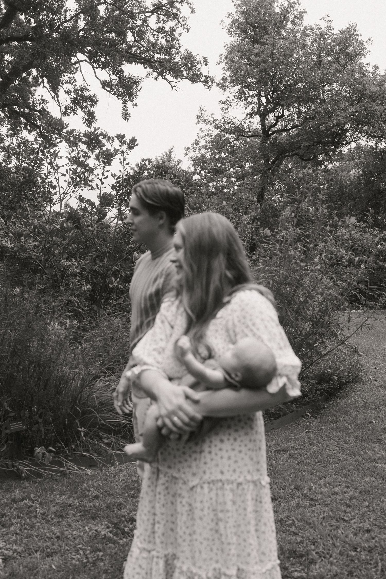 Black and white photo of a couple standing outdoors, woman cradling a newborn baby in a garden setting.