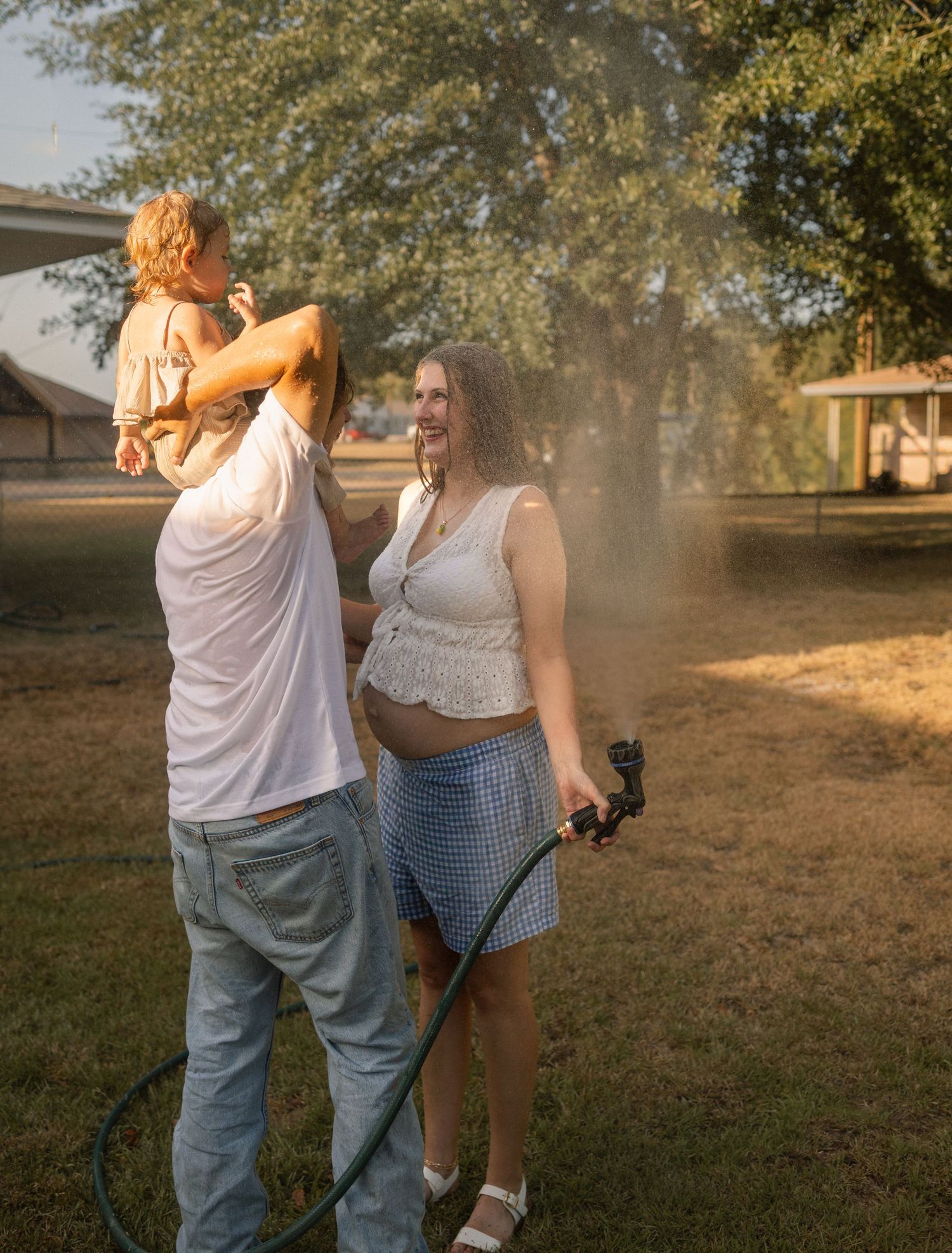 Pregnant woman holding garden hose laughing with partner carrying toddler in golden sunlight.