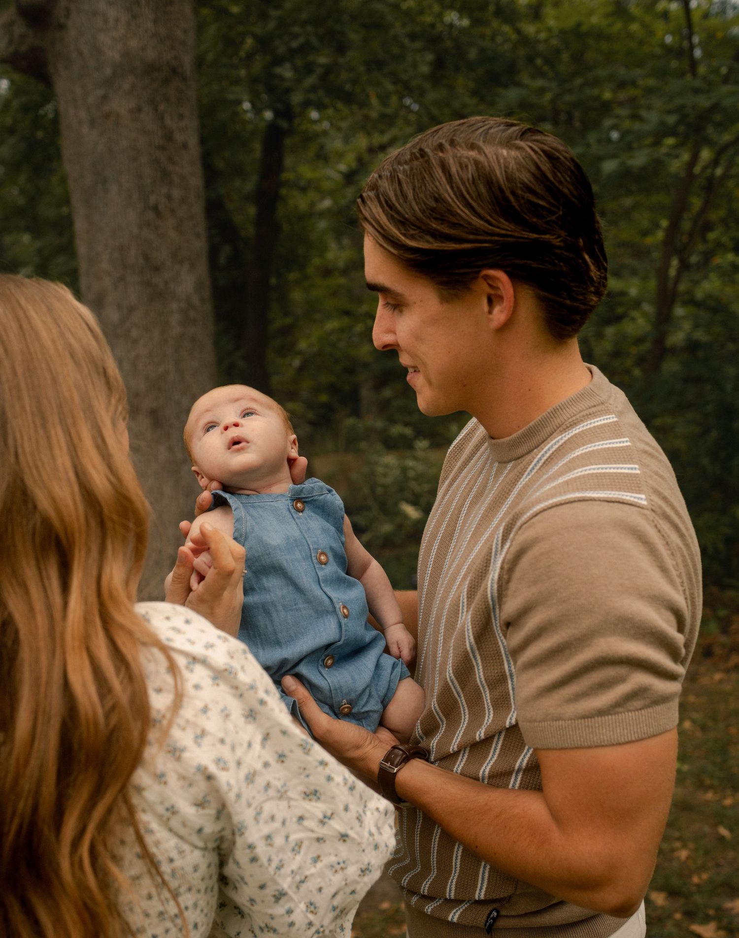 A young couple admiring their newborn baby outdoors surrounded by lush green trees.