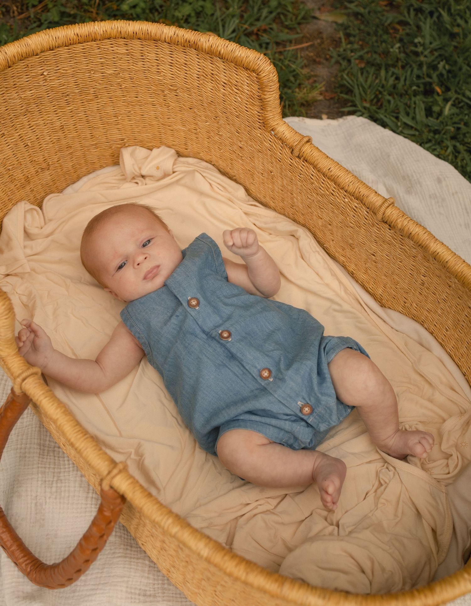 Newborn baby in blue romper lying in wicker moses basket on cream blanket outdoors.
