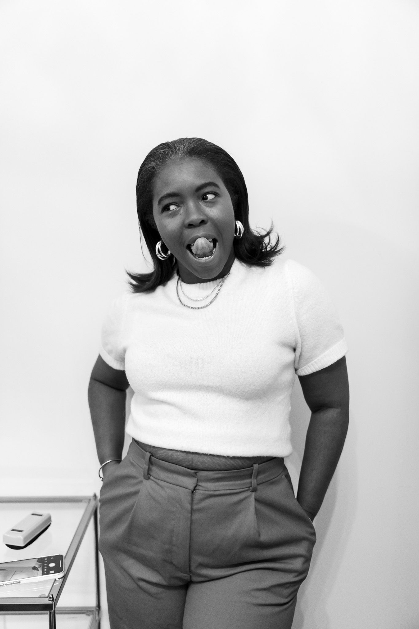 Black and white photo of a woman making a funny face with hands in pockets, wearing a white top and trousers.