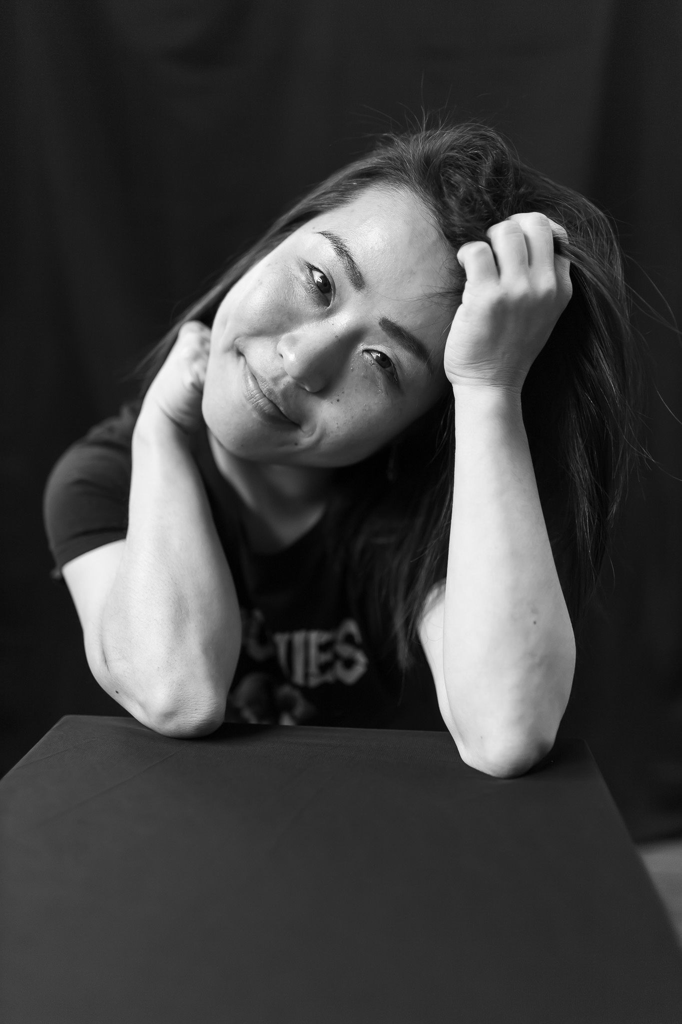 Black and white portrait of a woman with hands in her hair resting on a dark surface with a soft smile.