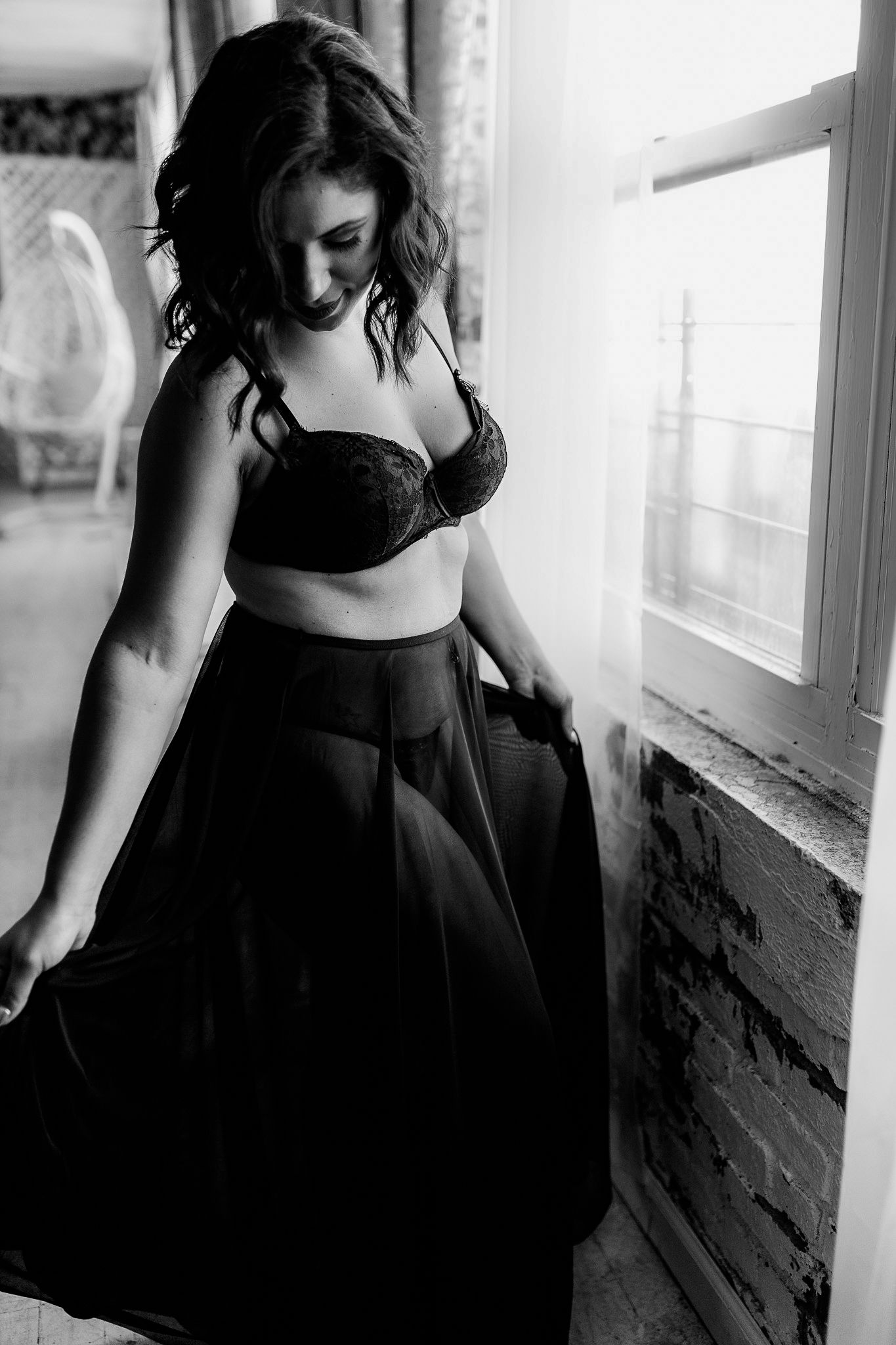 Elegant black and white boudoir photo of a woman in lingerie posing by a window.