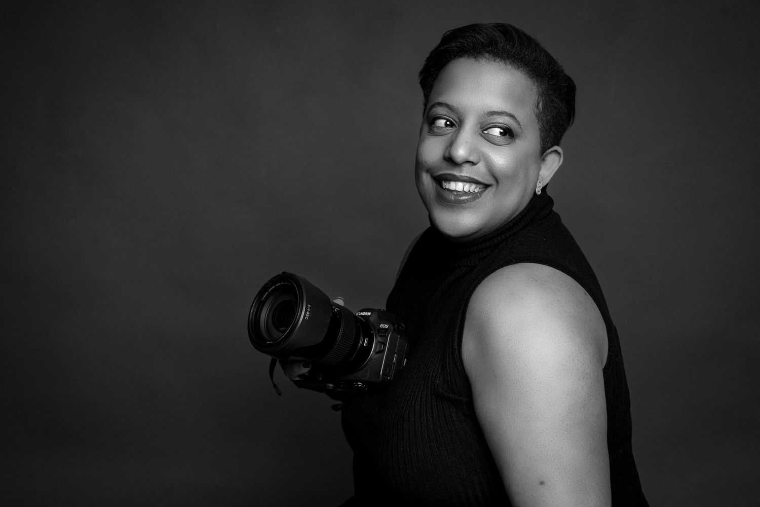 Black and white portrait of a smiling woman holding a professional camera at her side in a studio.