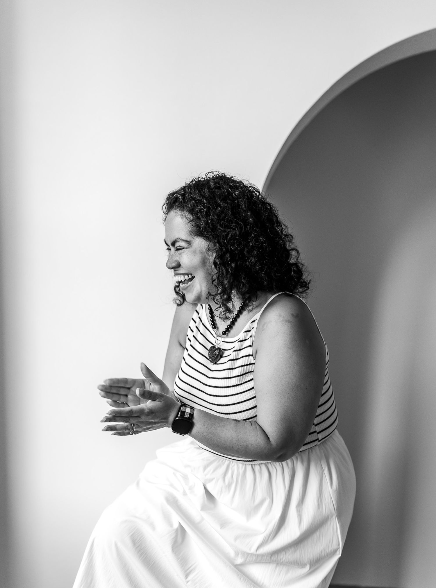 Black and white photo of a curly-haired woman laughing, wearing striped top and white skirt, seated near an arch.