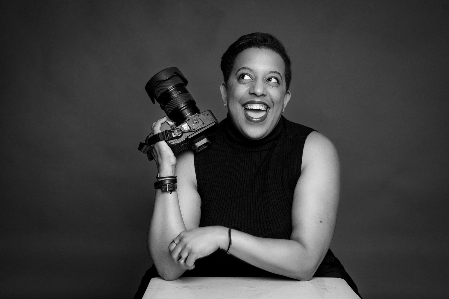 Black and white studio portrait of a laughing woman holding a camera while seated at a table.