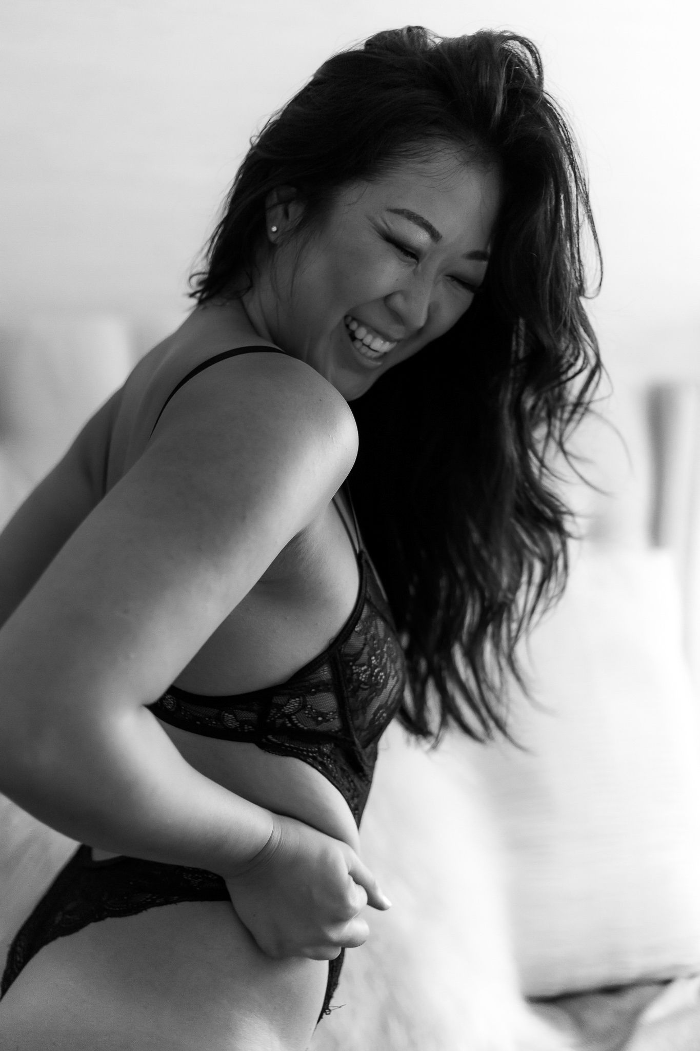 Black and white boudoir portrait of smiling woman in lace lingerie on bed.