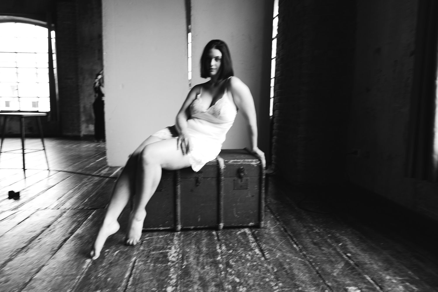 Black and white full-length boudoir photo of a woman on a vintage trunk by a window.