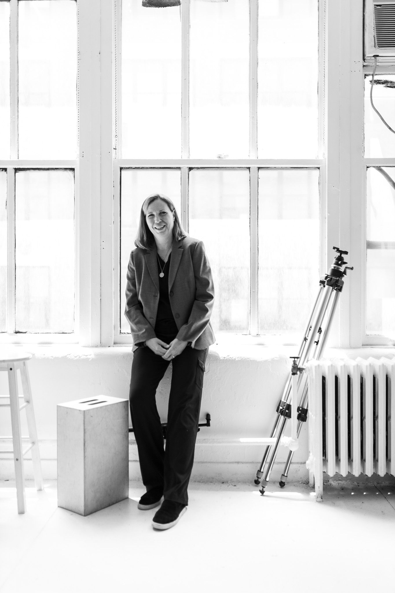 Woman in blazer smiling in bright studio with large windows and camera tripod.