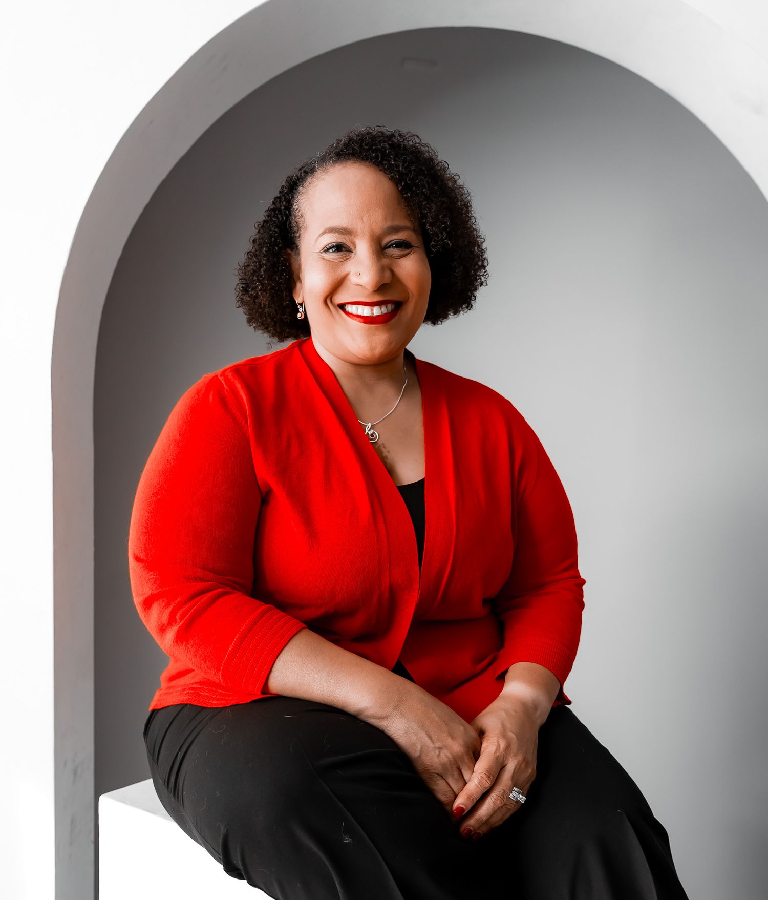 Smiling woman in red cardigan sitting confidently under a white arch in a professional portrait photo.