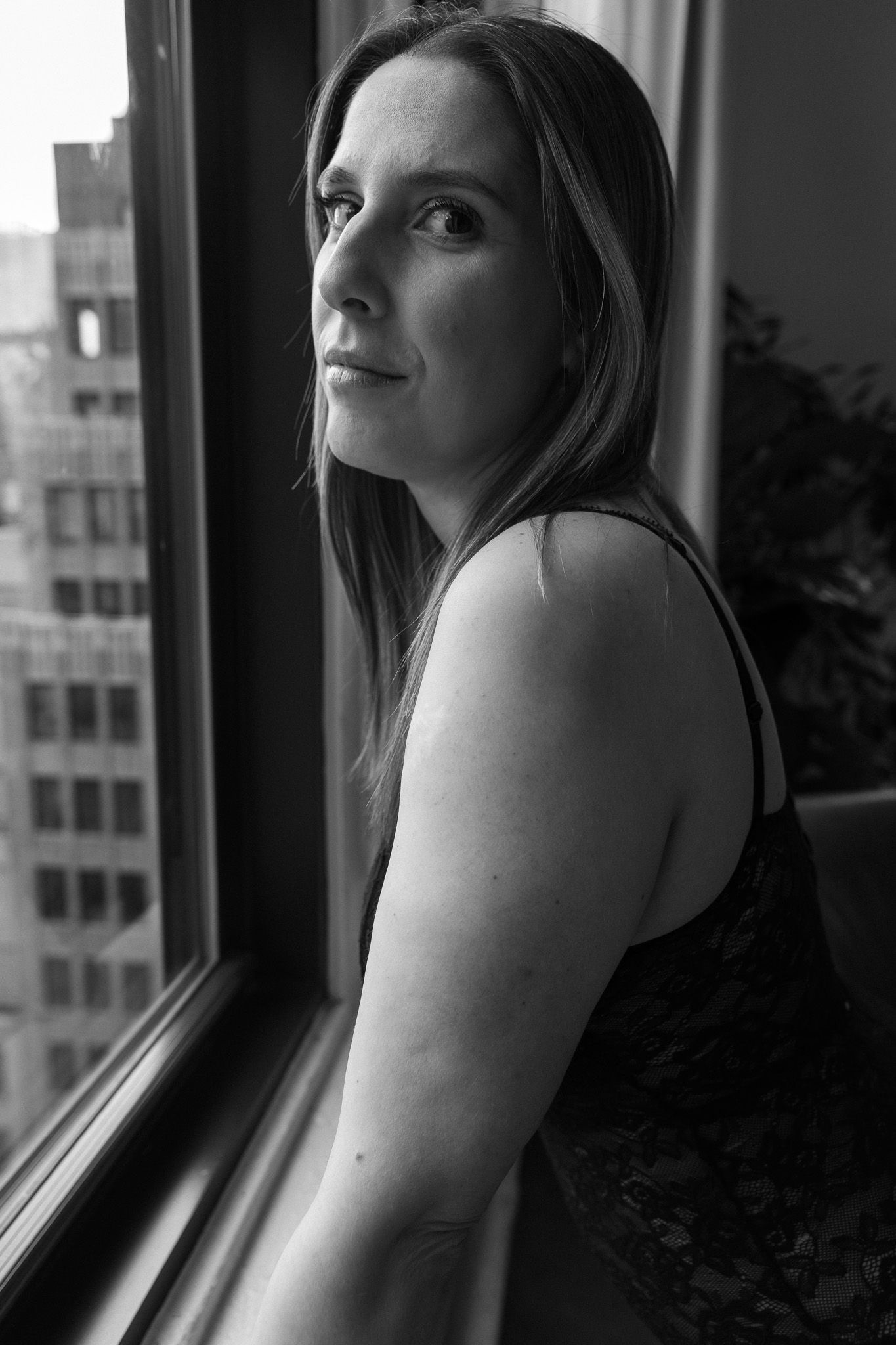 Black and white portrait of a woman in black lace top looking over her shoulder near a city-view window.