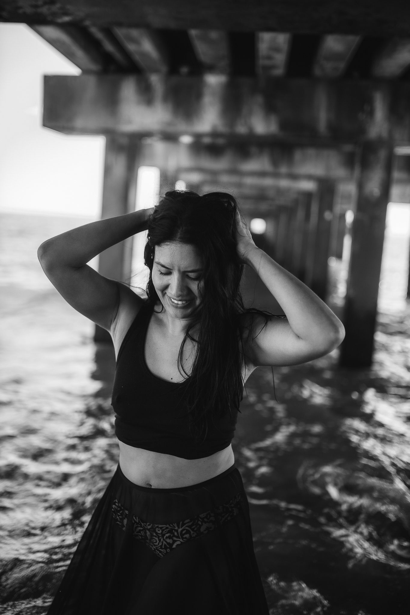 Black and white photo of a smiling woman in crop top under a pier by the ocean.