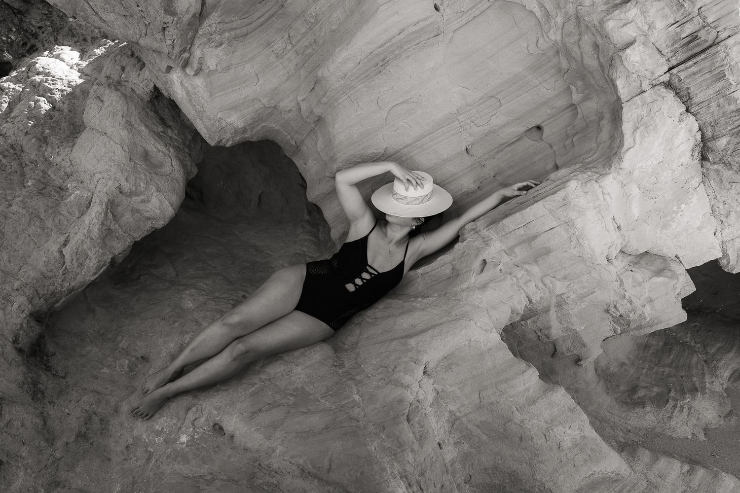 Aerial black and white photo of woman in black swimsuit and hat reclining gracefully among desert rock formations.