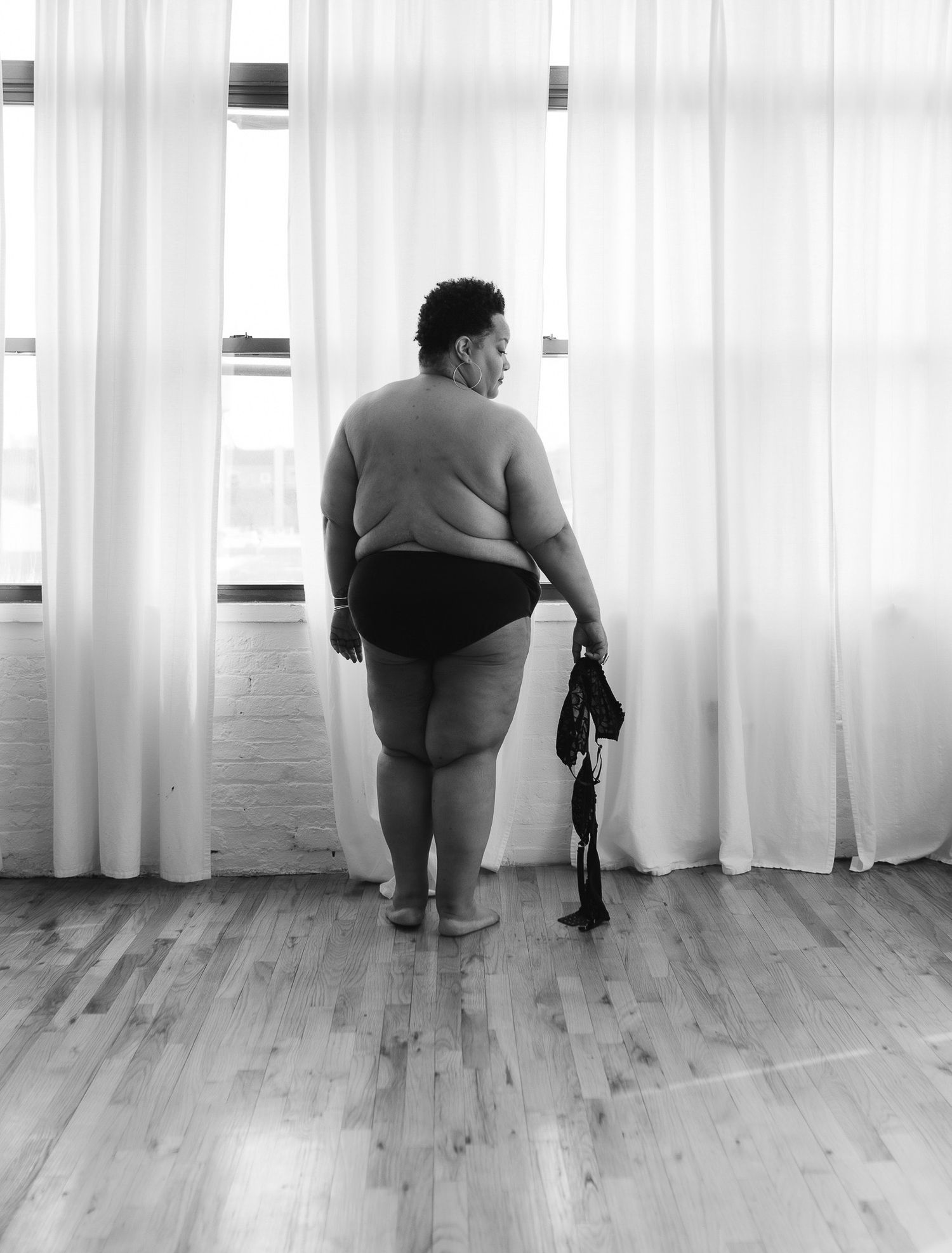 Plus-size woman standing in underwear facing curtained windows in black and white dance studio photo.