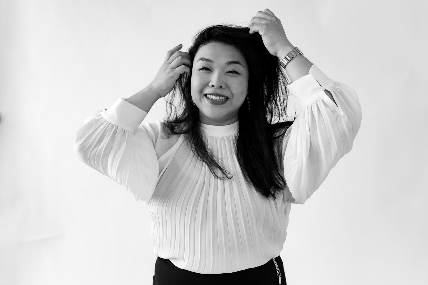 Smiling Asian woman in white pleated blouse fixing hair against white background in black and white.
