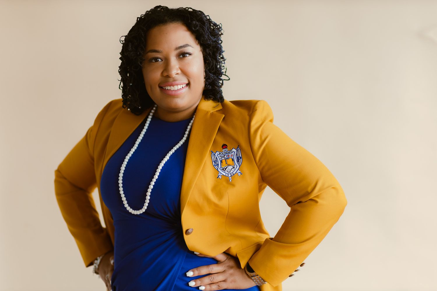 Woman in mustard yellow blazer and blue dress with pearl necklace and brooch, smiling confidently.