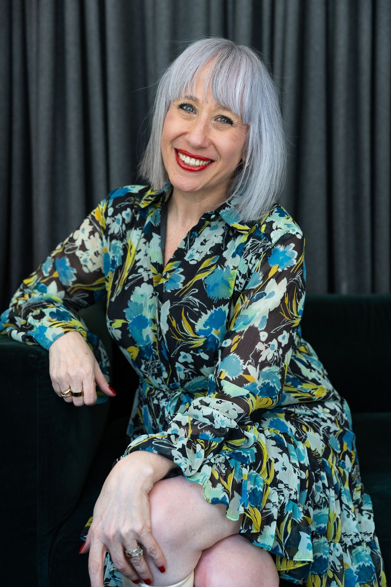 Smiling woman with silver hair wearing a colorful floral dress, seated on a dark sofa against a gray curtain backdrop.