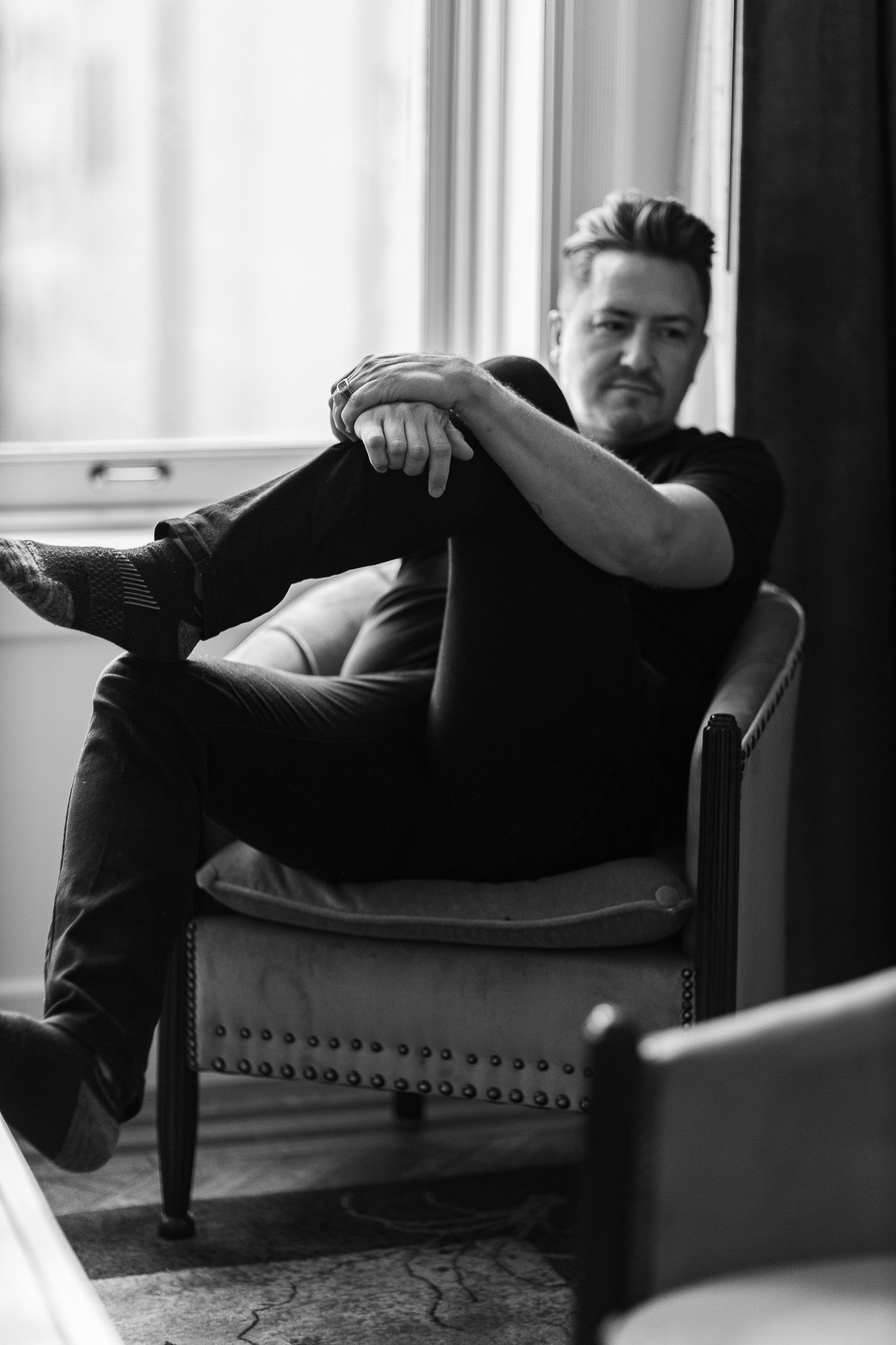 Man in all black outfit sitting in vintage armchair by window, looking at camera.