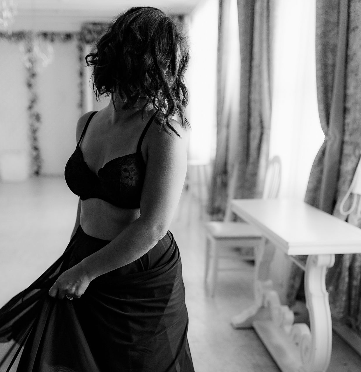 Black and white photo of a woman in lingerie holding a dark skirt in a bright room.