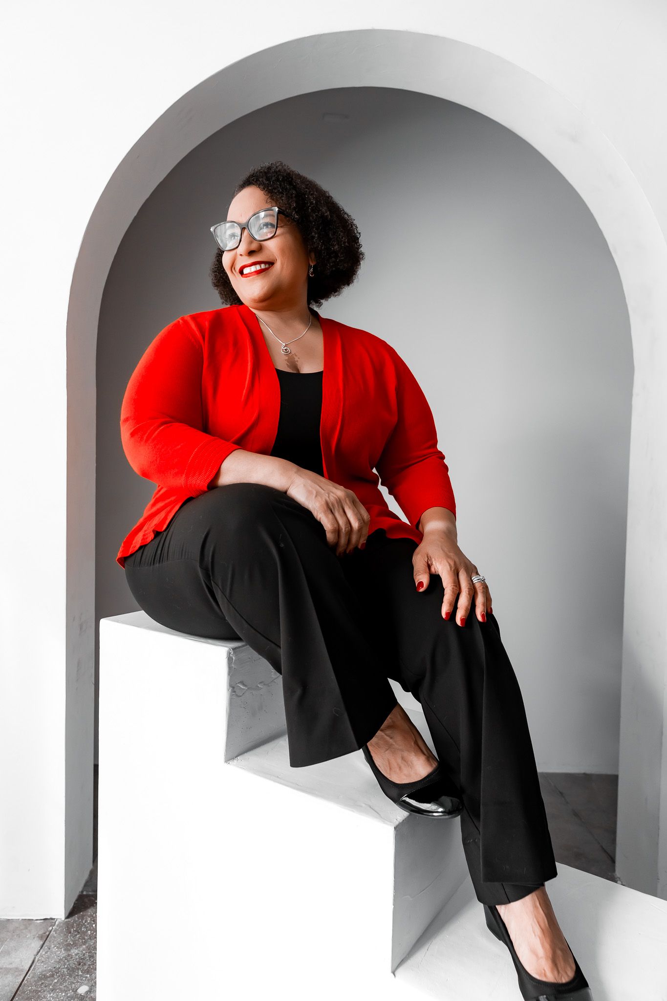 Woman in red cardigan and black pants sitting in an arched alcove, smiling confidently, wearing glasses.