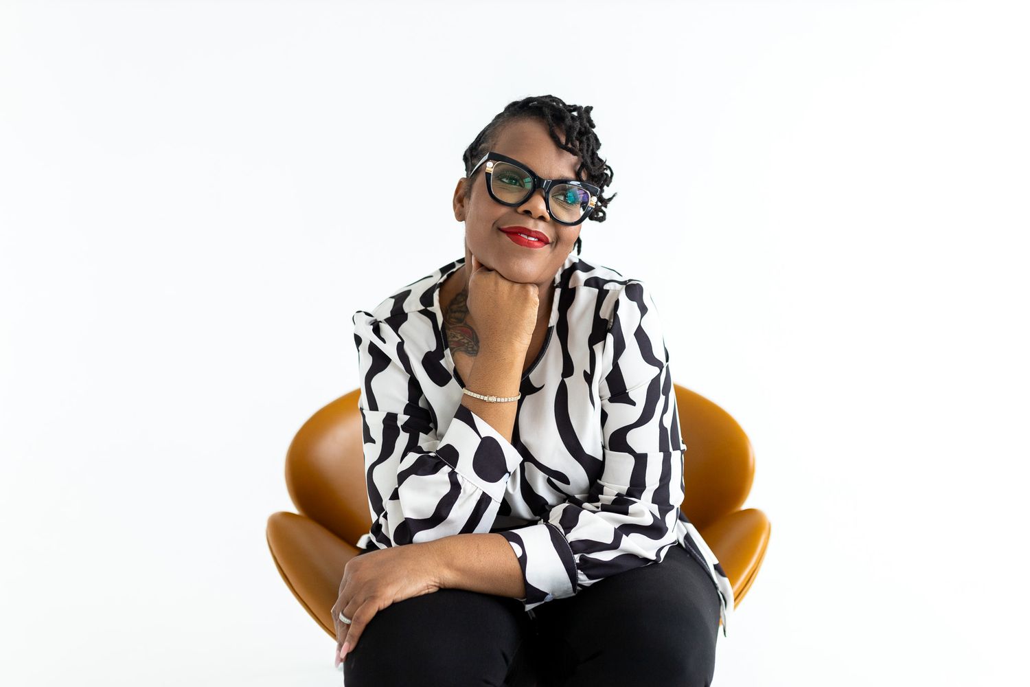 Woman in black and white patterned shirt sitting in orange chair, wearing green glasses, posing confidently.