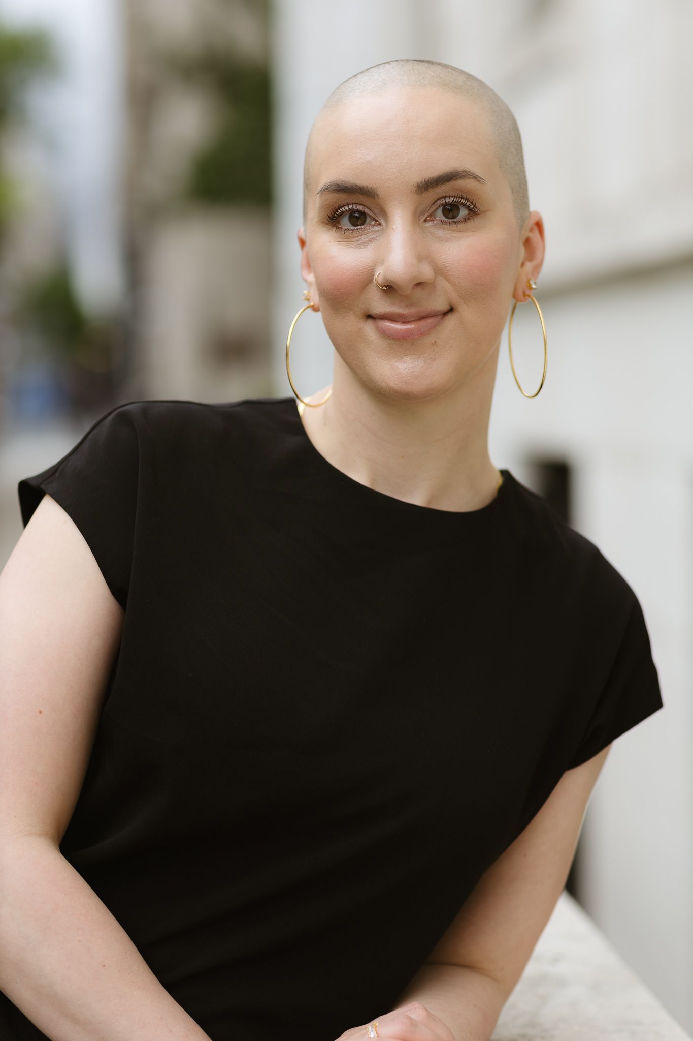 Woman with shaved head wearing gold hoop earrings and black sleeveless top, smiling outdoors.