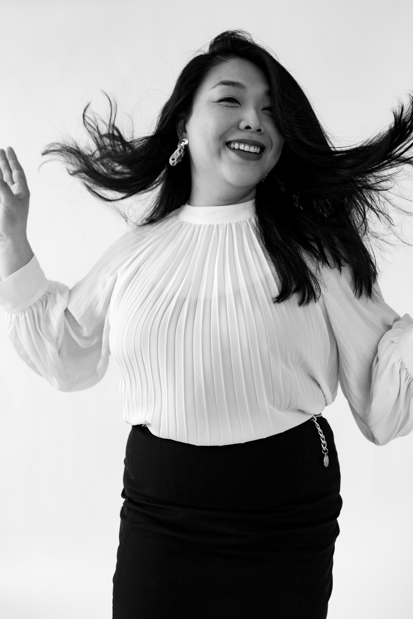 Black and white photo of a joyful Asian woman in white pleated blouse and black skirt, hair flowing, arms raised.