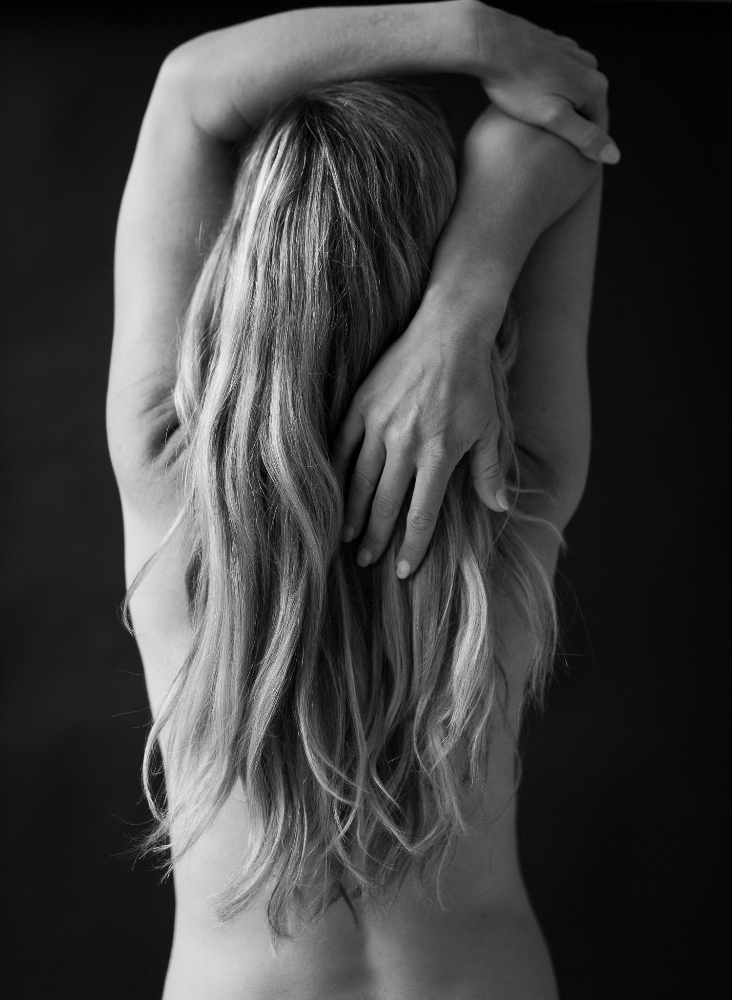 Black and white photo of a woman with long blonde hair, arms raised, seen from behind.