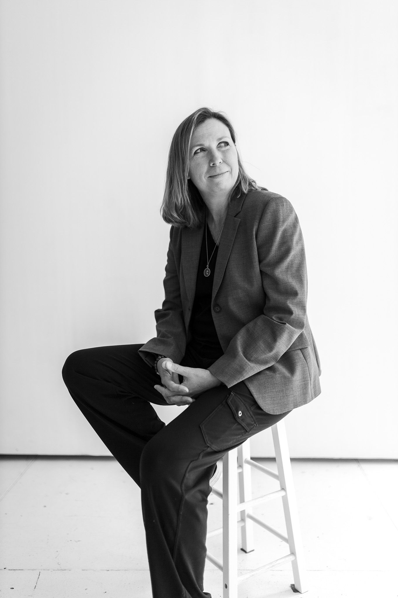 Woman in gray blazer sitting on stool with hands clasped gazing upward in studio.