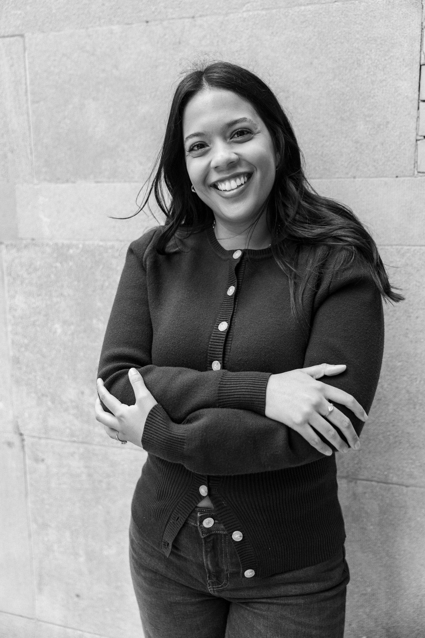 Black and white portrait of a smiling young woman with crossed arms leaning against a stone wall.