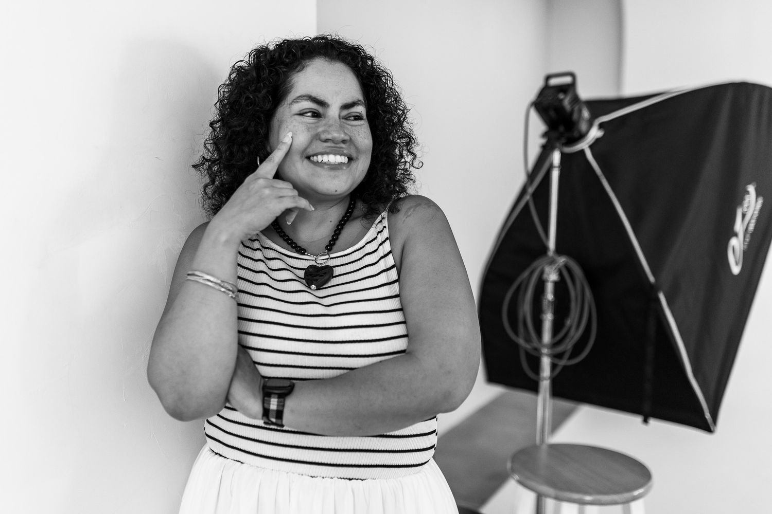 Black and white photo of a smiling woman in striped top posing in a photography studio with lighting equipment.