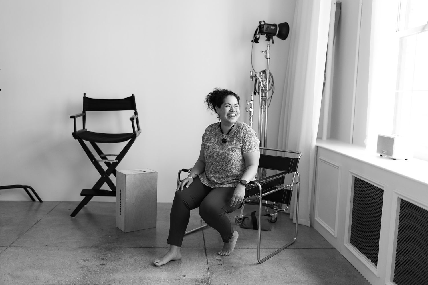 Black and white photo of a laughing woman sitting in a chair in a photography studio with equipment.