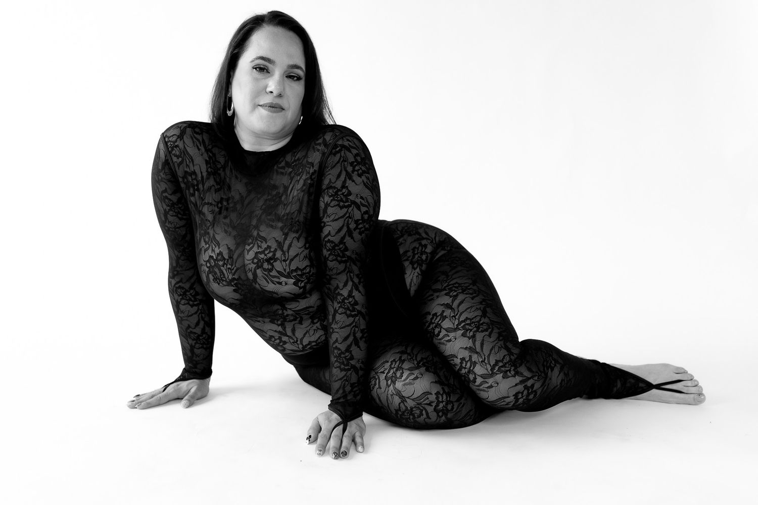 Woman in black lace bodysuit posing gracefully on white studio floor in black and white photography.