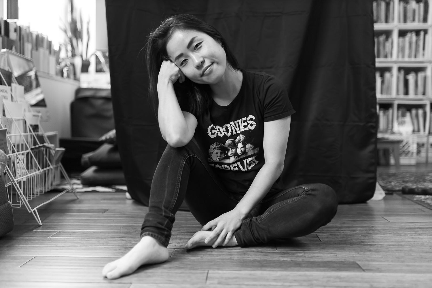 Asian woman sitting barefoot on wooden floor, resting head on hand, wearing Goonies t-shirt, contemplative black and white photo.