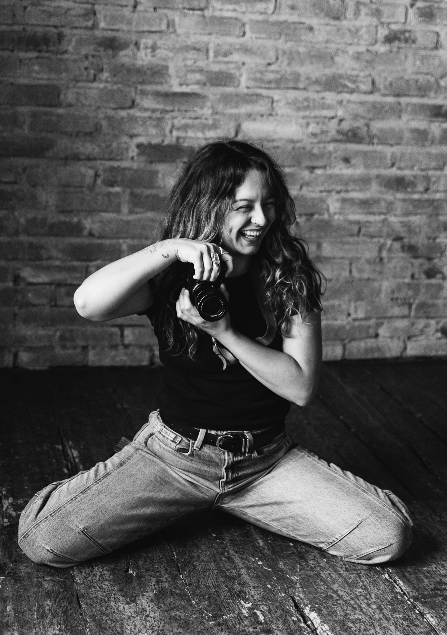 Black and white photo of a smiling woman sitting on floor holding a camera against a brick wall.