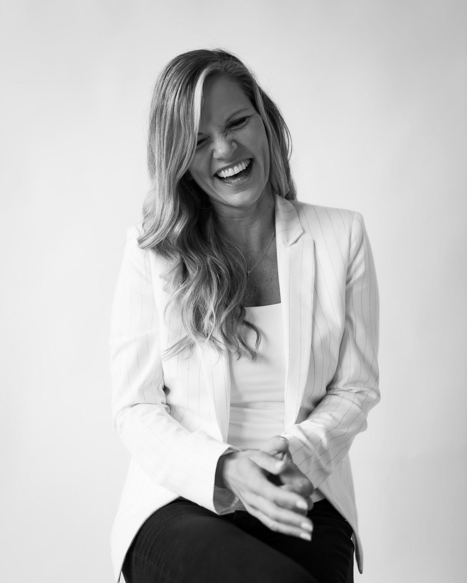 Smiling blonde woman in white blazer laughing joyfully in a black and white studio portrait.