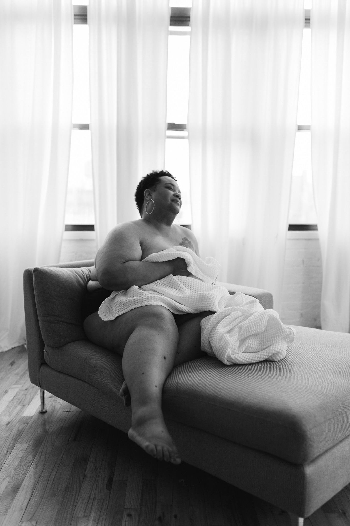 Black and white boudoir photo of a plus-size woman wrapped in a white robe relaxing on a chaise lounge by sheer curtains.