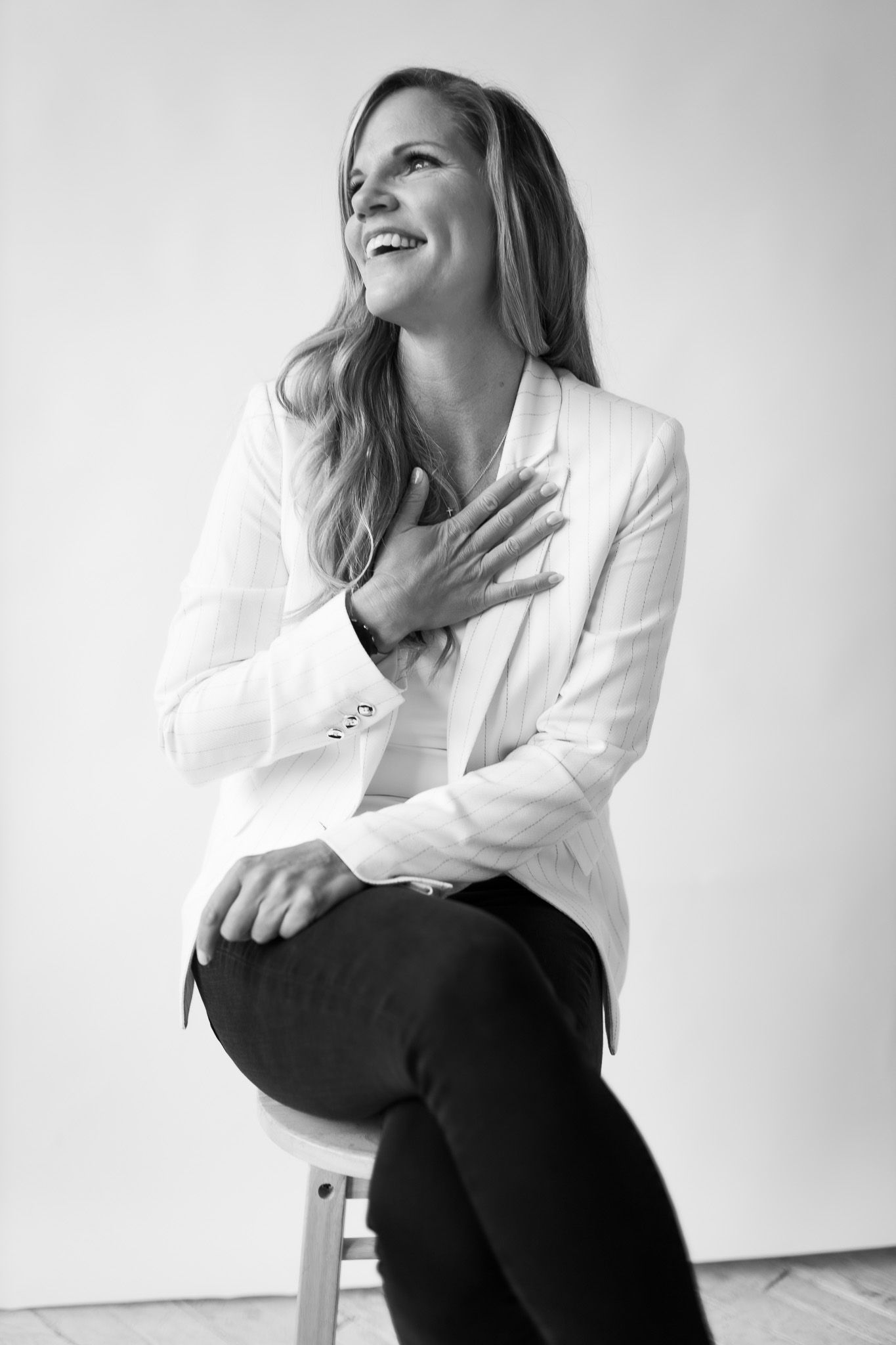 Woman in white blazer sits smiling with hand on chest in elegant black and white portrait photo.