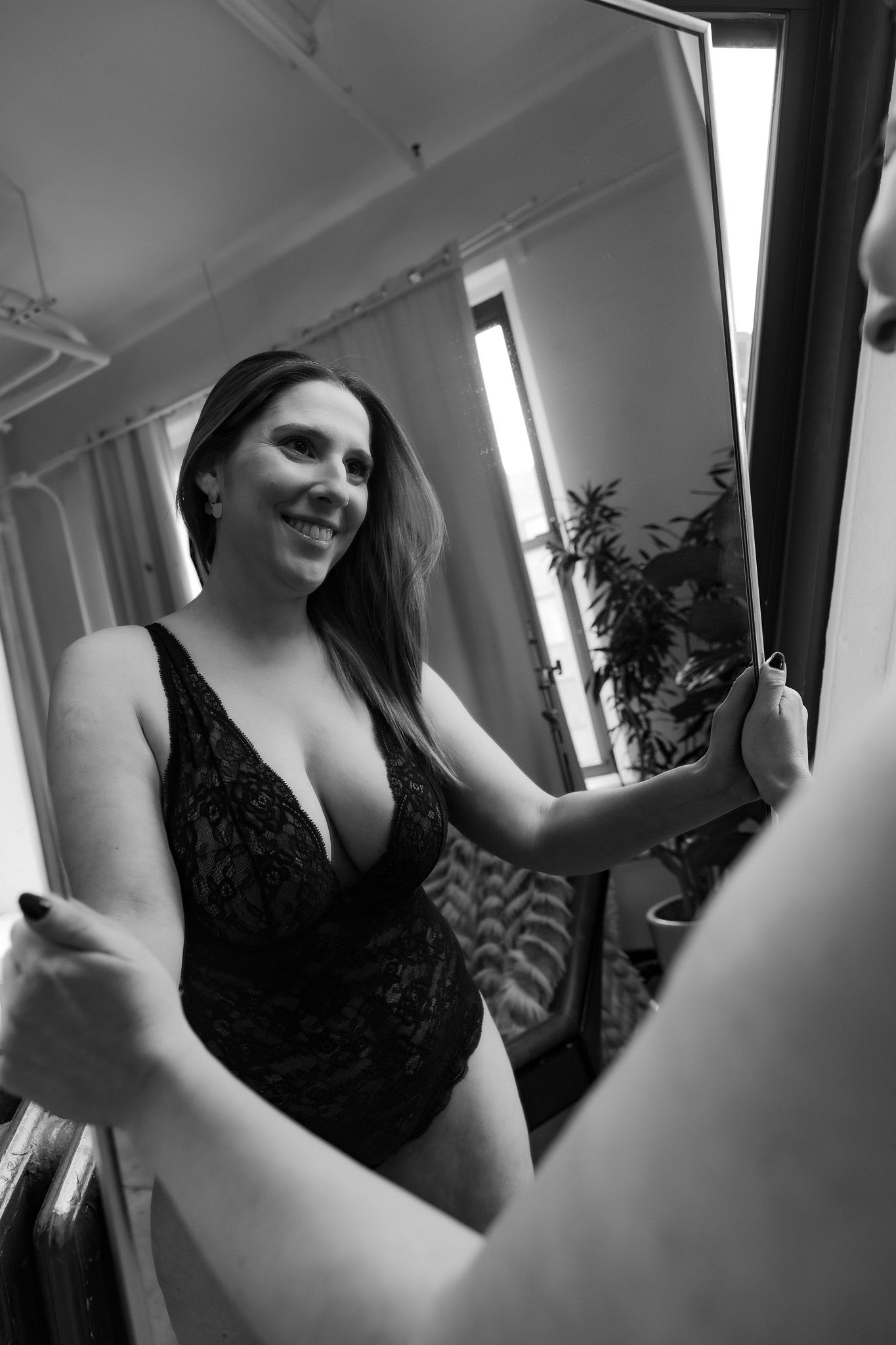 Black and white boudoir photo of a woman in black lace bodysuit smiling at her reflection in a mirror.